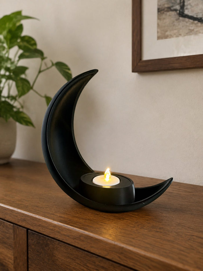 Charcoal black 3D printed crescent moon votive holder with a lit LED tea light, on a wooden sideboard beside a pothos plant